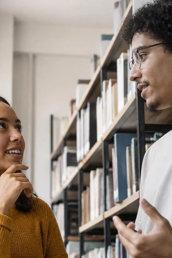Duas participantes de pé conversando no corredor entre prateleiras de livros